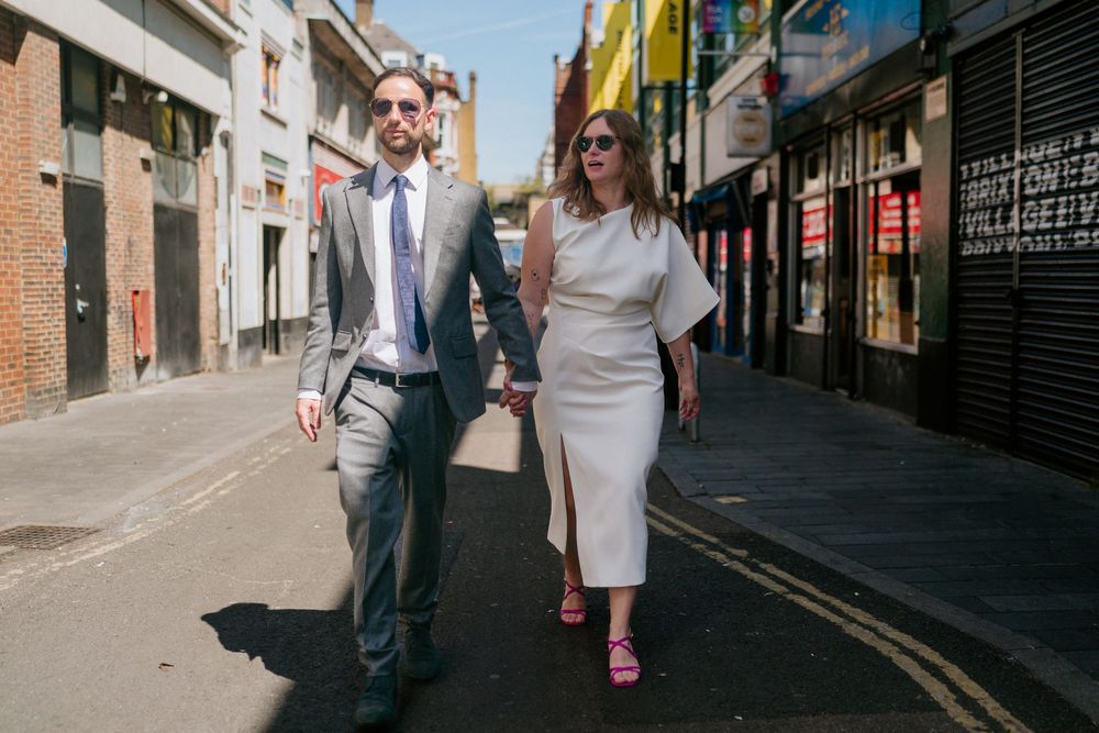 Couple walking hand in hand down a sunny urban street, man in grey suit, woman in white dress with pink heels.