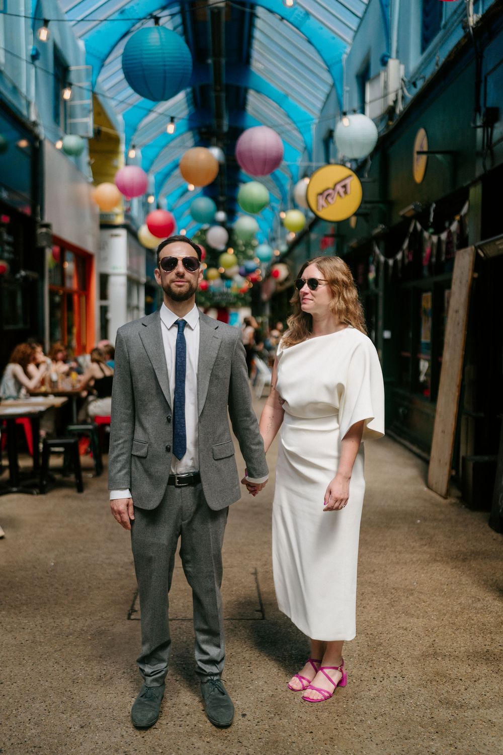 A couple in wedding attire holds hands under colorful paper lanterns in a vibrant indoor market arcade.