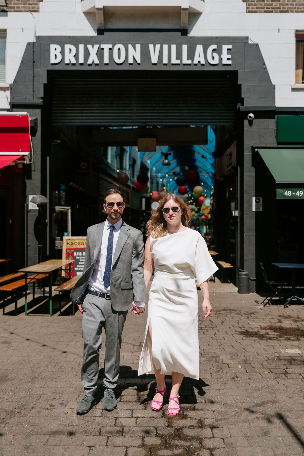 Couple holding hands in front of Brixton Village market, bride in white dress with pink shoes, groom in grey suit.