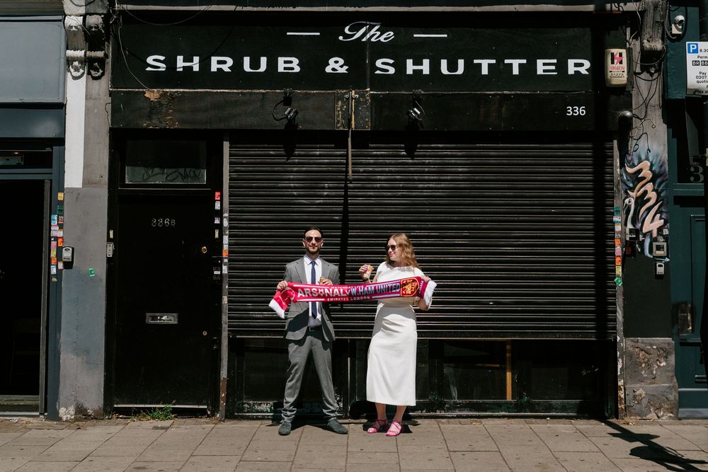 Couple holding a red scarf outside The Shrub & Shutter bar in London, dressed in wedding attire.