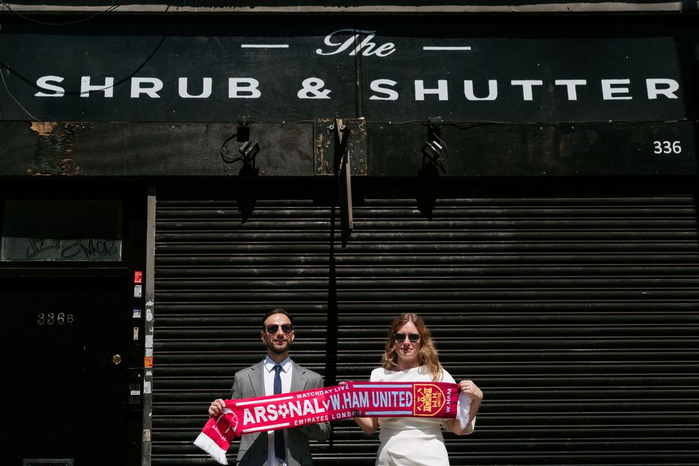 Couple holding Arsenal and West Ham United scarves outside The Shrub & Shutter bar on their wedding day.