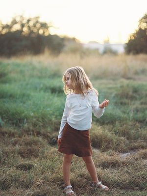 Child in white top and brown skirt dancing in sunlit field at golden hour.