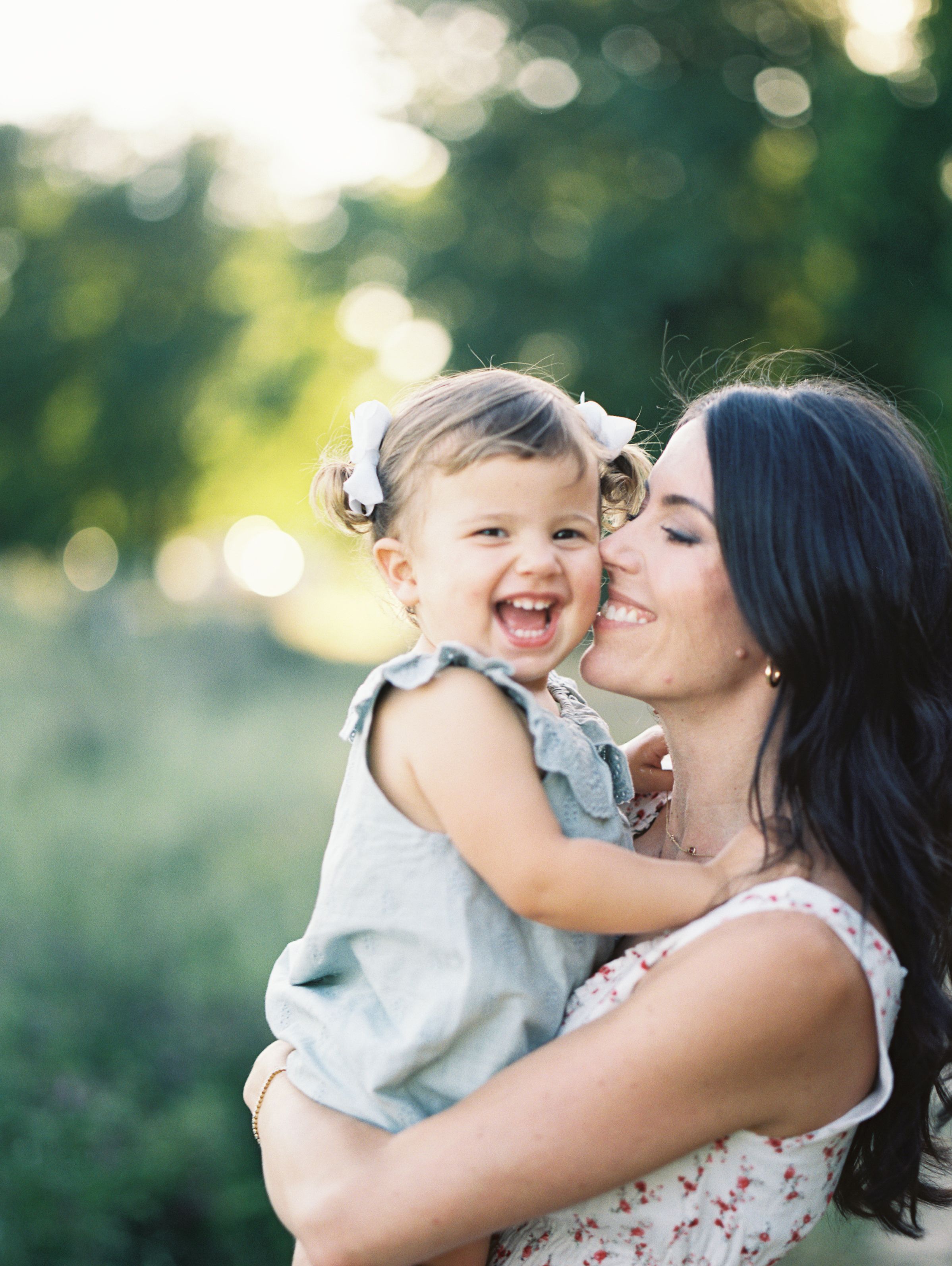 A joyful toddler girl laughs while being held by her dark-haired mother outdoors in a sunlit green field.