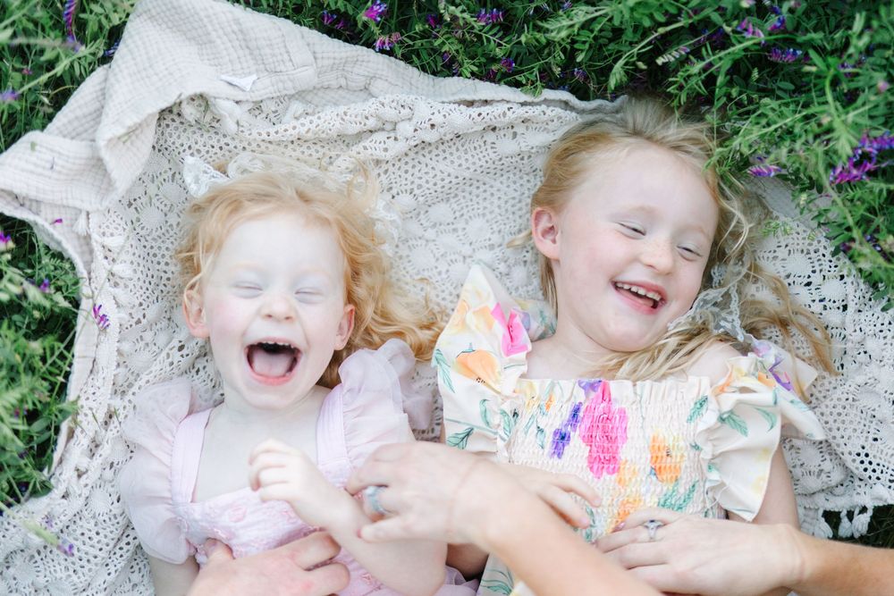 Two laughing girls with curly red hair lying on a lace blanket surrounded by purple flowers outdoors.