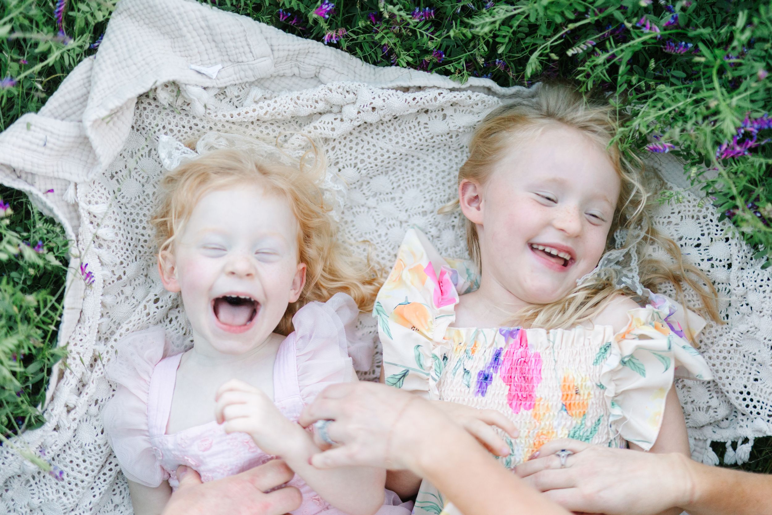 Two laughing girls with curly red hair lying on a lace blanket surrounded by purple flowers outdoors.