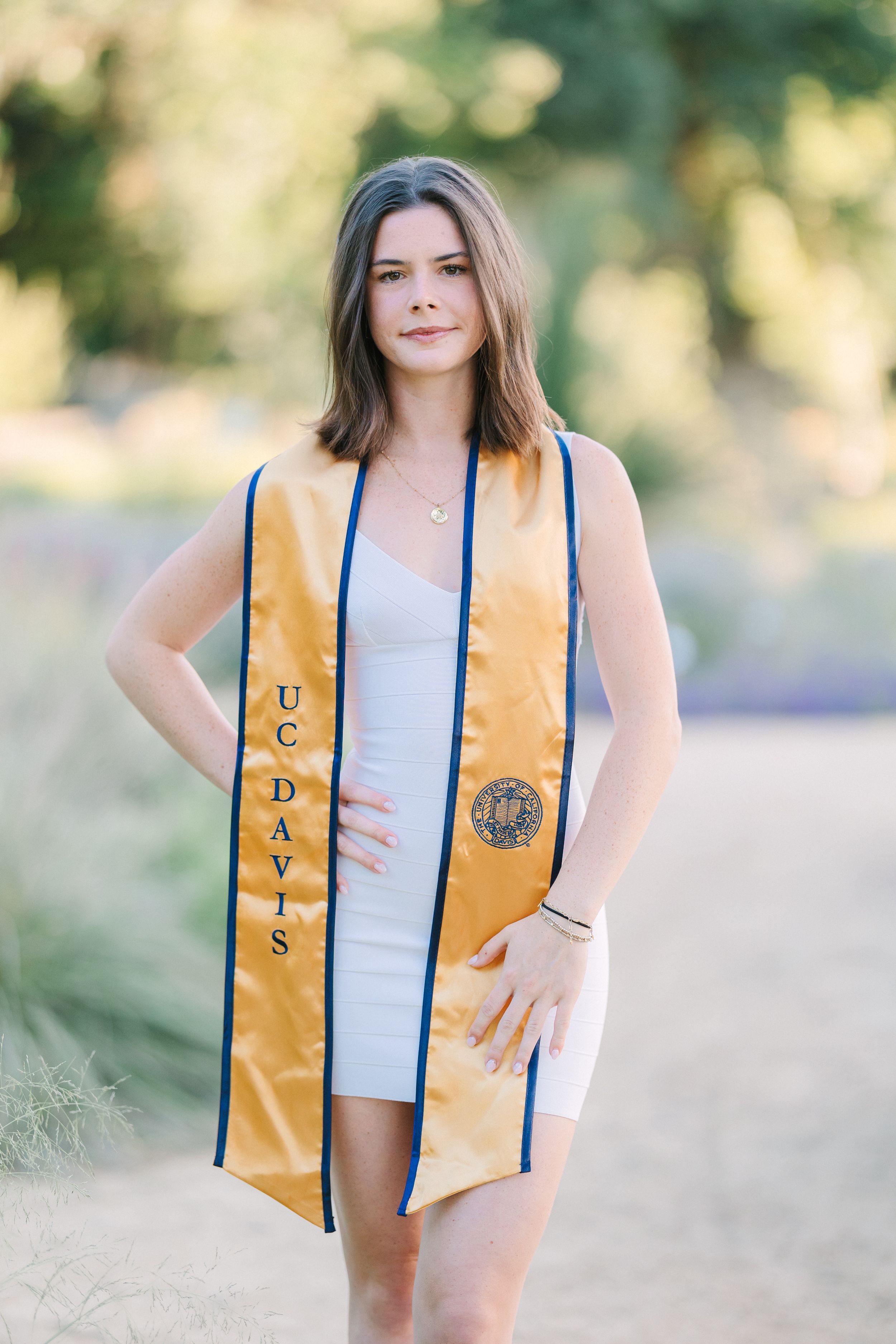 UC Davis graduate in gold sash poses outdoors in white dress for senior graduation portrait photo.