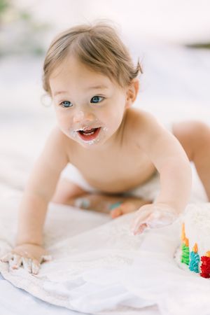 Smiling baby in diaper crawling toward a small birthday cake with colorful candles, face covered in frosting.