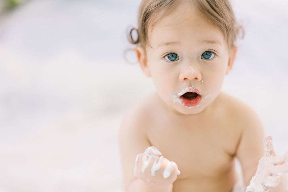 Adorable blue-eyed baby with cream on face and hands, looking surprised in a bright, airy setting.