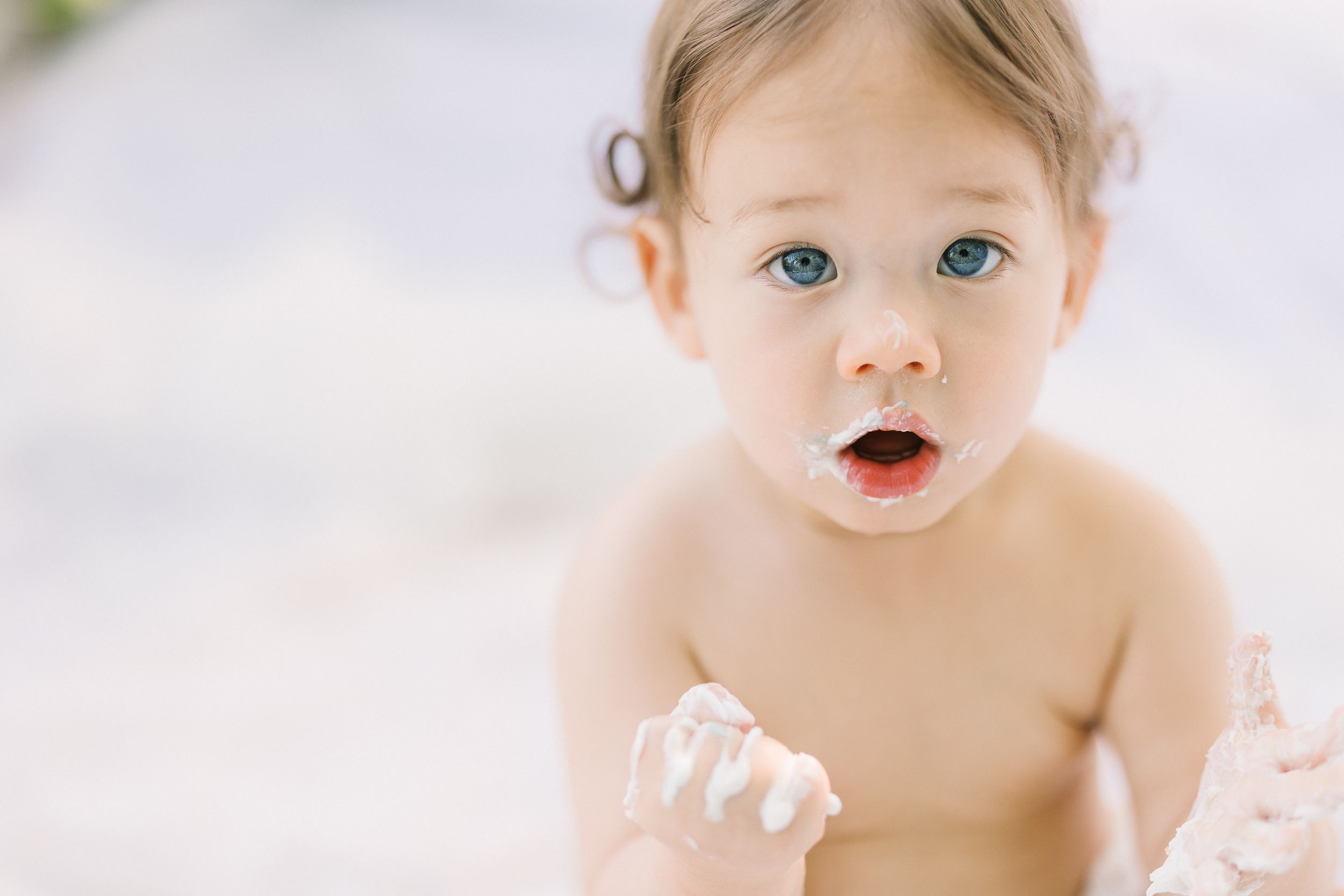 Adorable blue-eyed baby with cream on face and hands, looking surprised in a bright, airy setting.