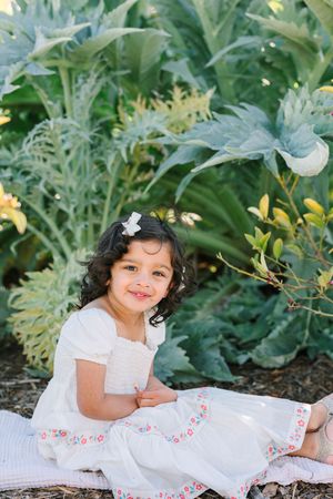 A child in a white dress with floral embroidery sits on a blanket in a garden.