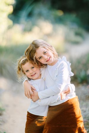 Two children in white tops and brown bottoms embracing while smiling at camera.