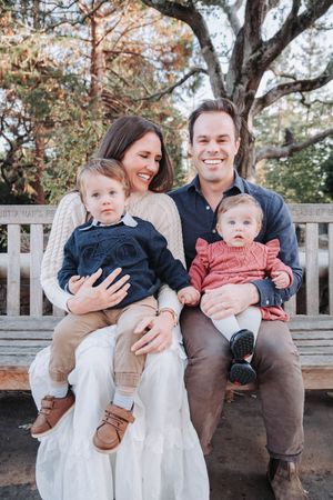 A family sits together on a wooden bench in a park during autumn, sharing playful moments with their young child.