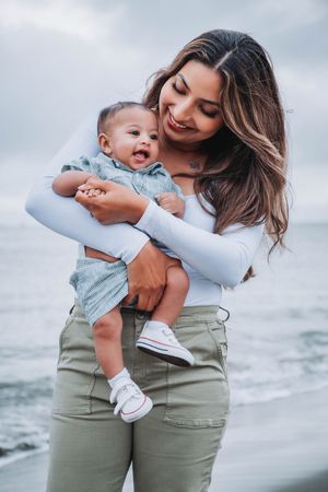 A tender moment between parent and baby in matching blue and olive outfits against a cloudy coastal backdrop.