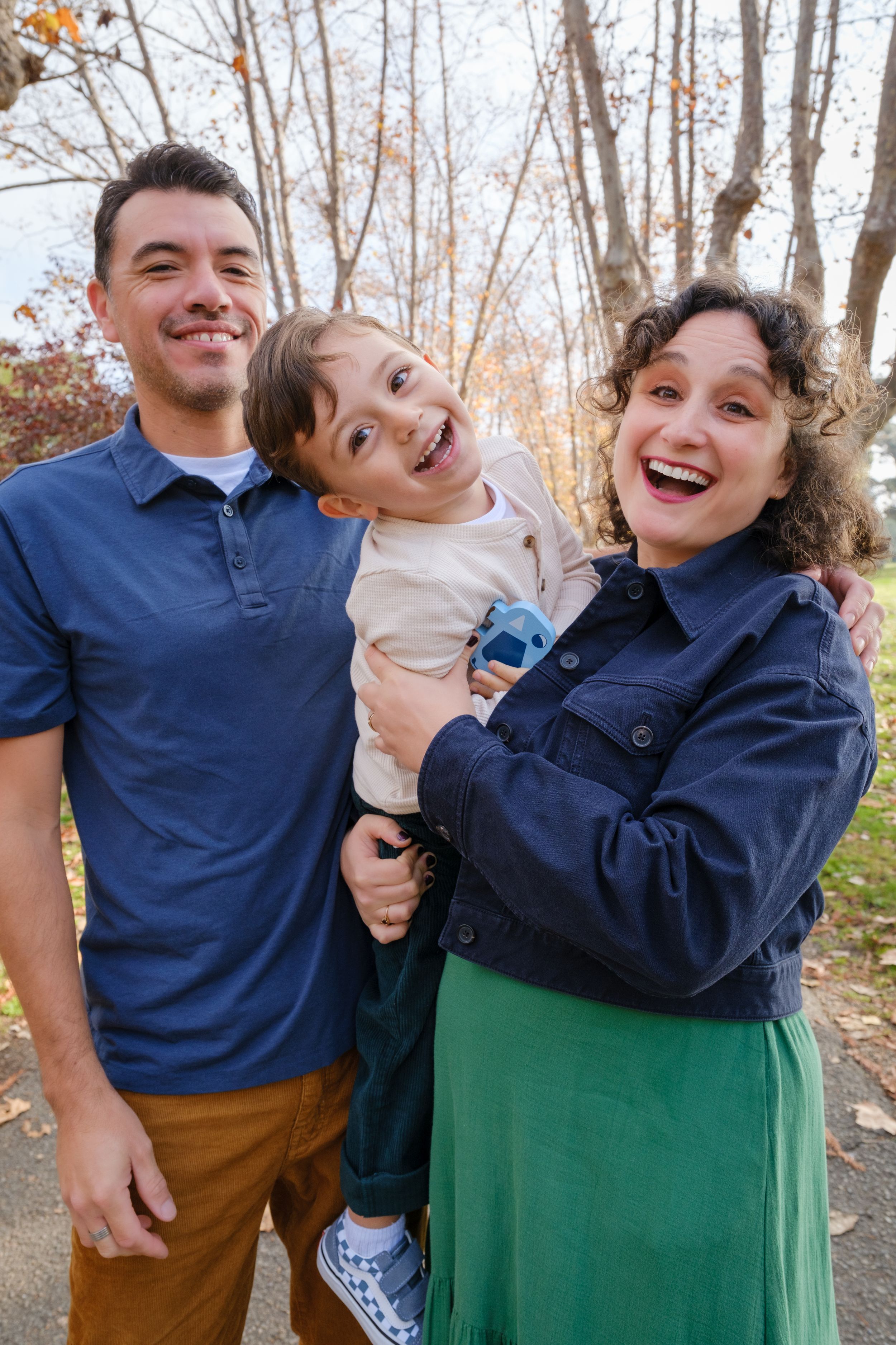 A happy family of three poses together outdoors on an autumn day with bare trees in the background.