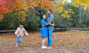 A couple embraces while standing on fallen autumn leaves as a child plays nearby in a vibrant fall setting.