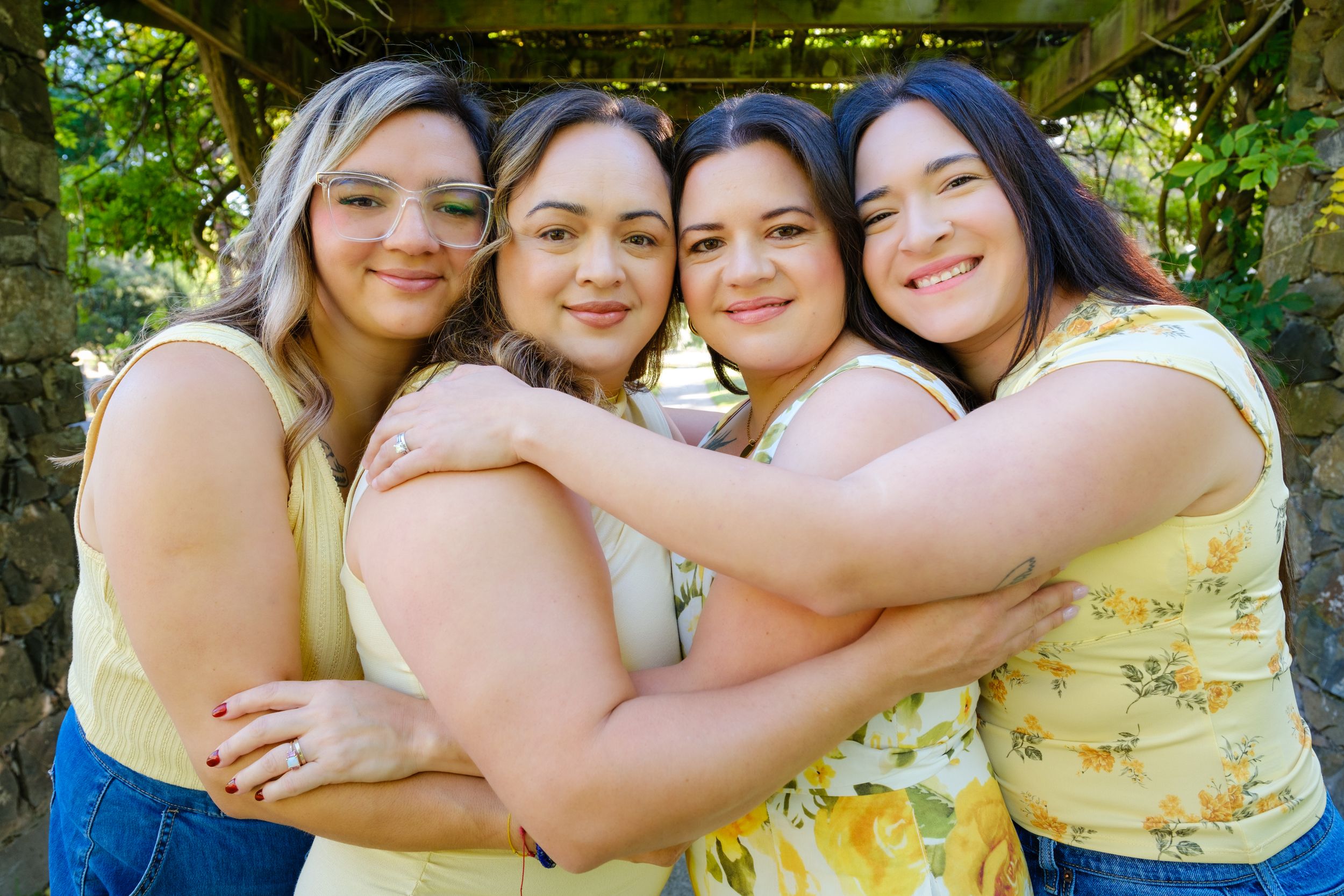 Four young women in yellow tops hugging and smiling outdoors under a wooden arbor with green foliage.