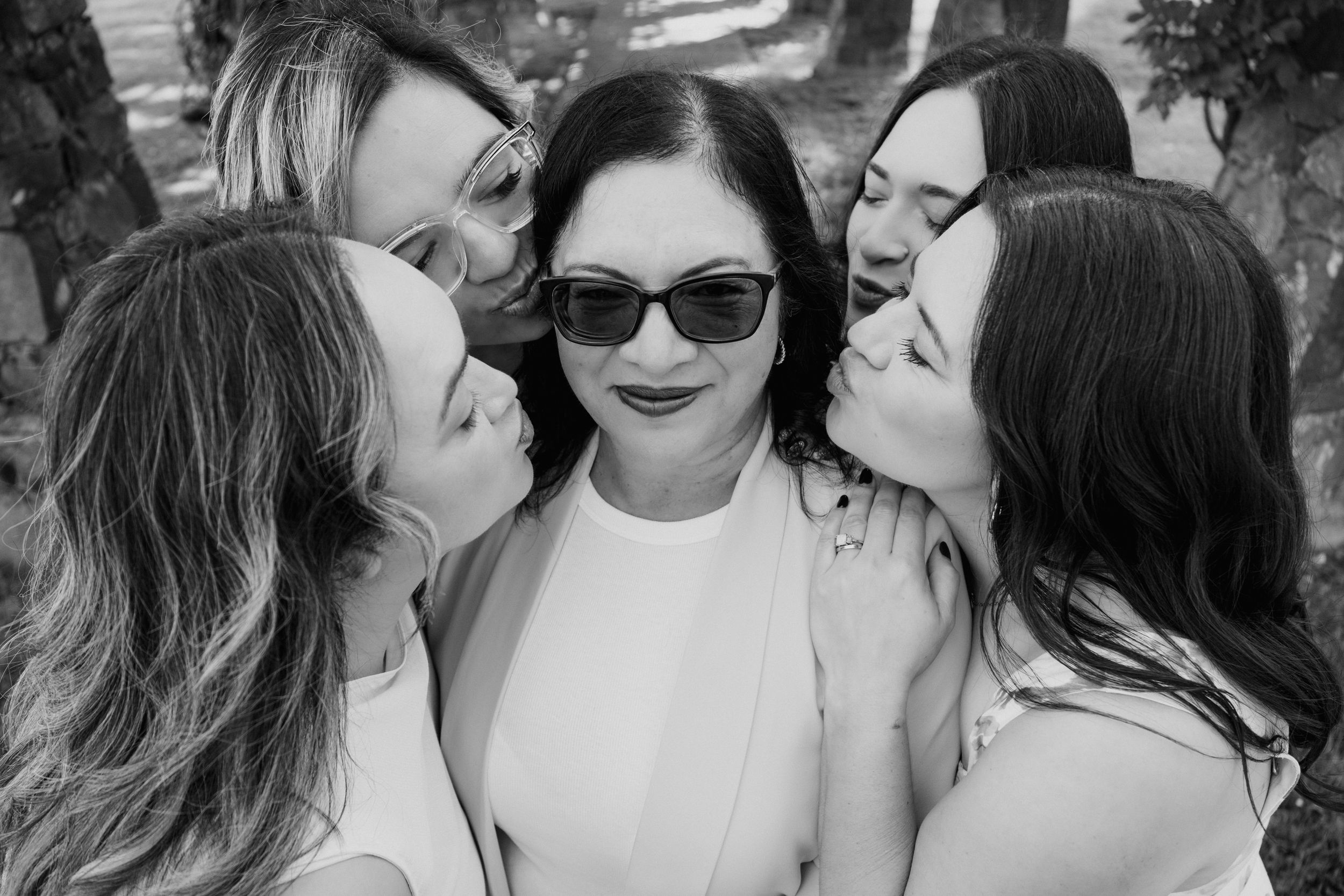 Black and white photo of five women, four leaning in to kiss one woman wearing sunglasses and white dress.
