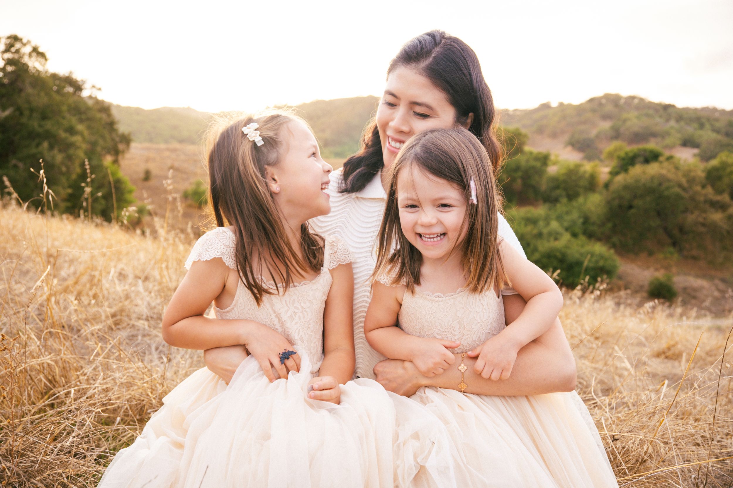 Three people in white dresses share a joyful moment together in a golden sunlit field during sunset.