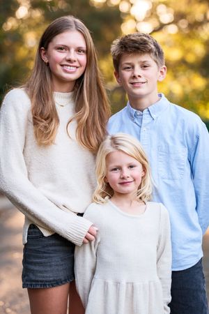Three siblings in coordinating white and blue outfits pose together during a golden hour outdoor photo session.