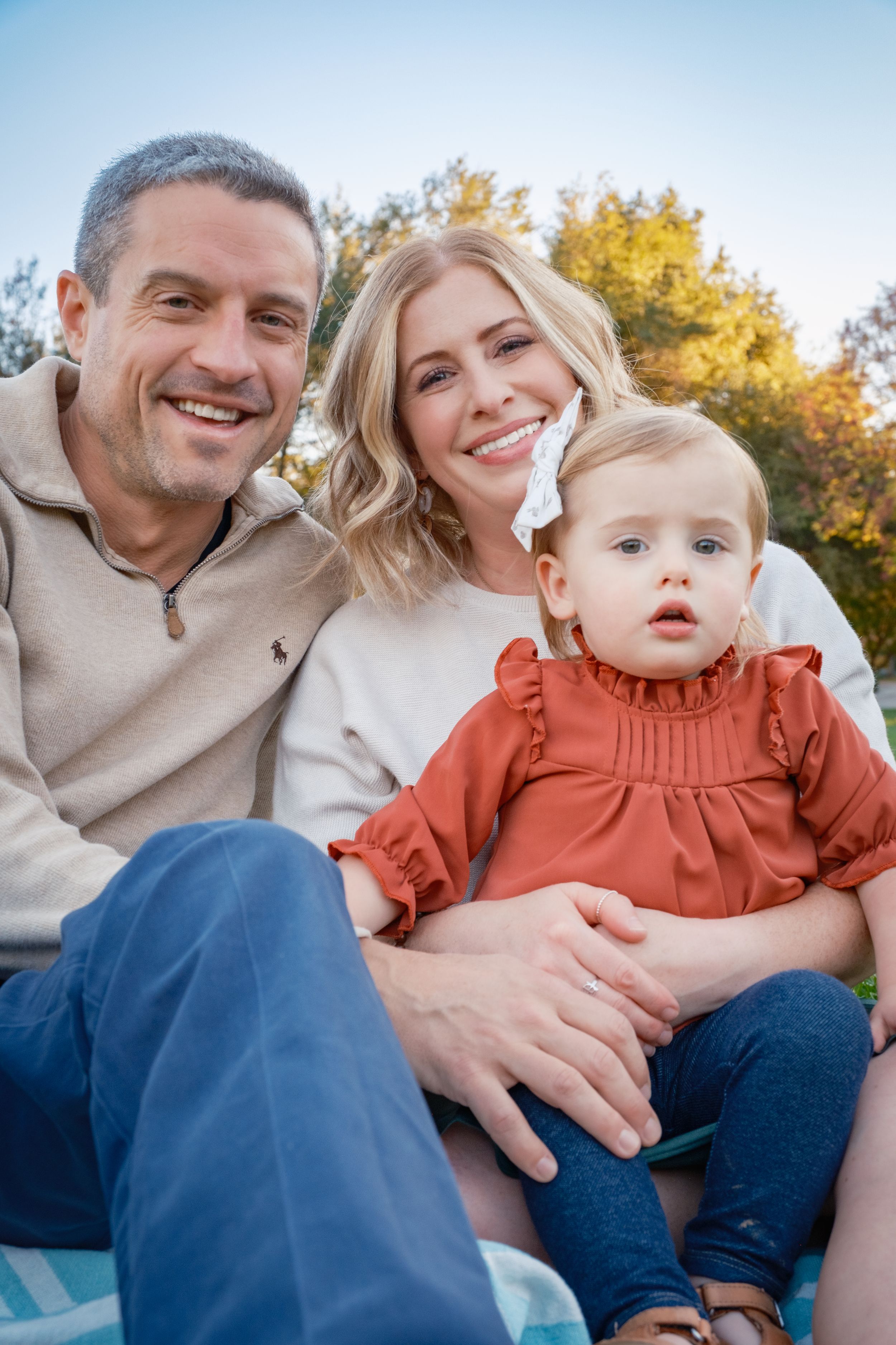 A family sits together outdoors during autumn, sharing warm smiles against a blue sky background.