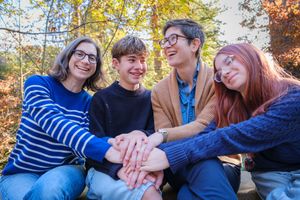 A group of friends stack their hands together in unity while sitting outdoors on a sunny autumn day.