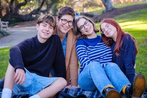 A group of four friends wearing casual clothing sit together outdoors on a sunny autumn day in a park setting.