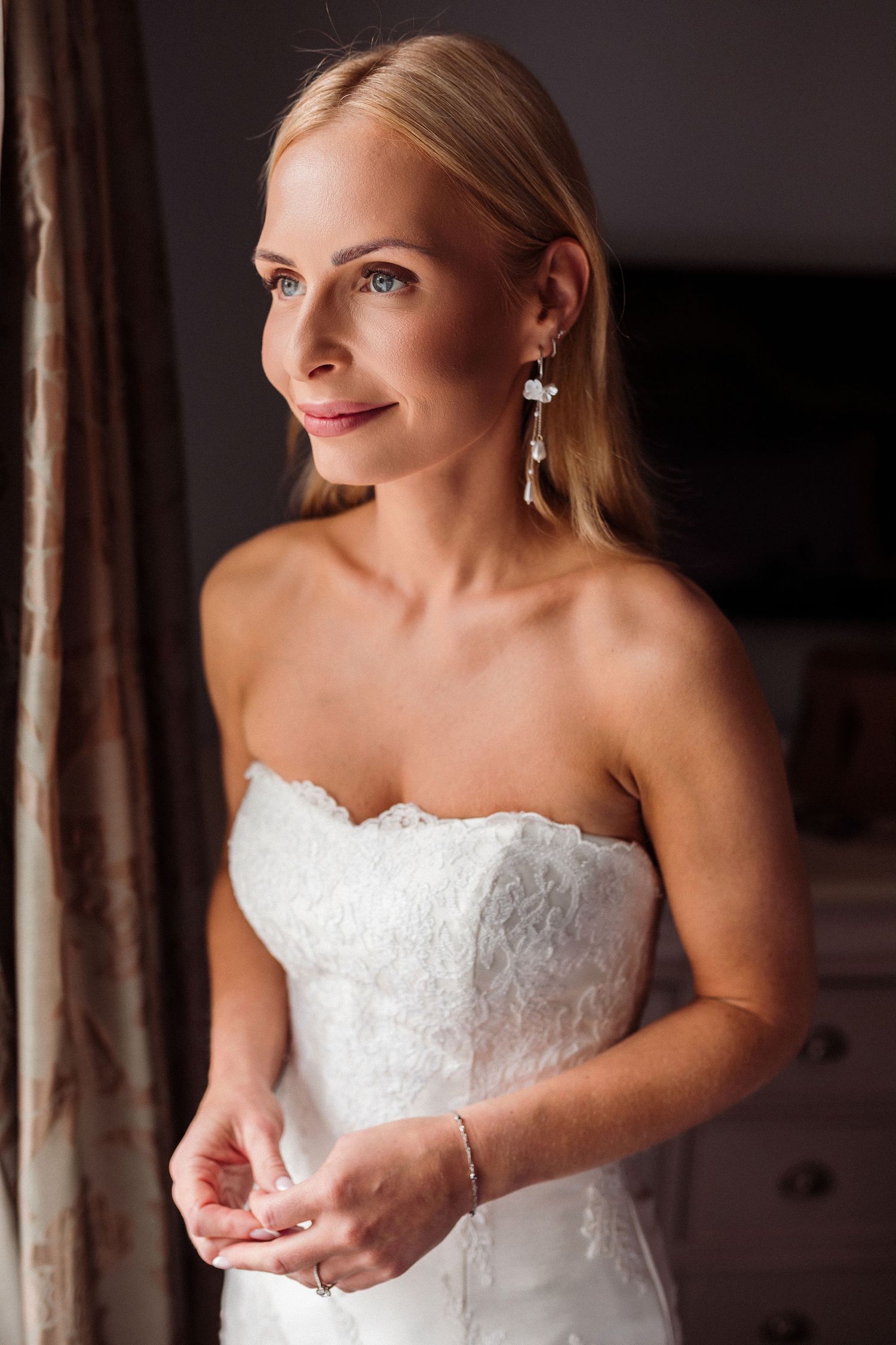 A bride in a white strapless wedding dress stands in soft lighting with elegant dangling earrings.