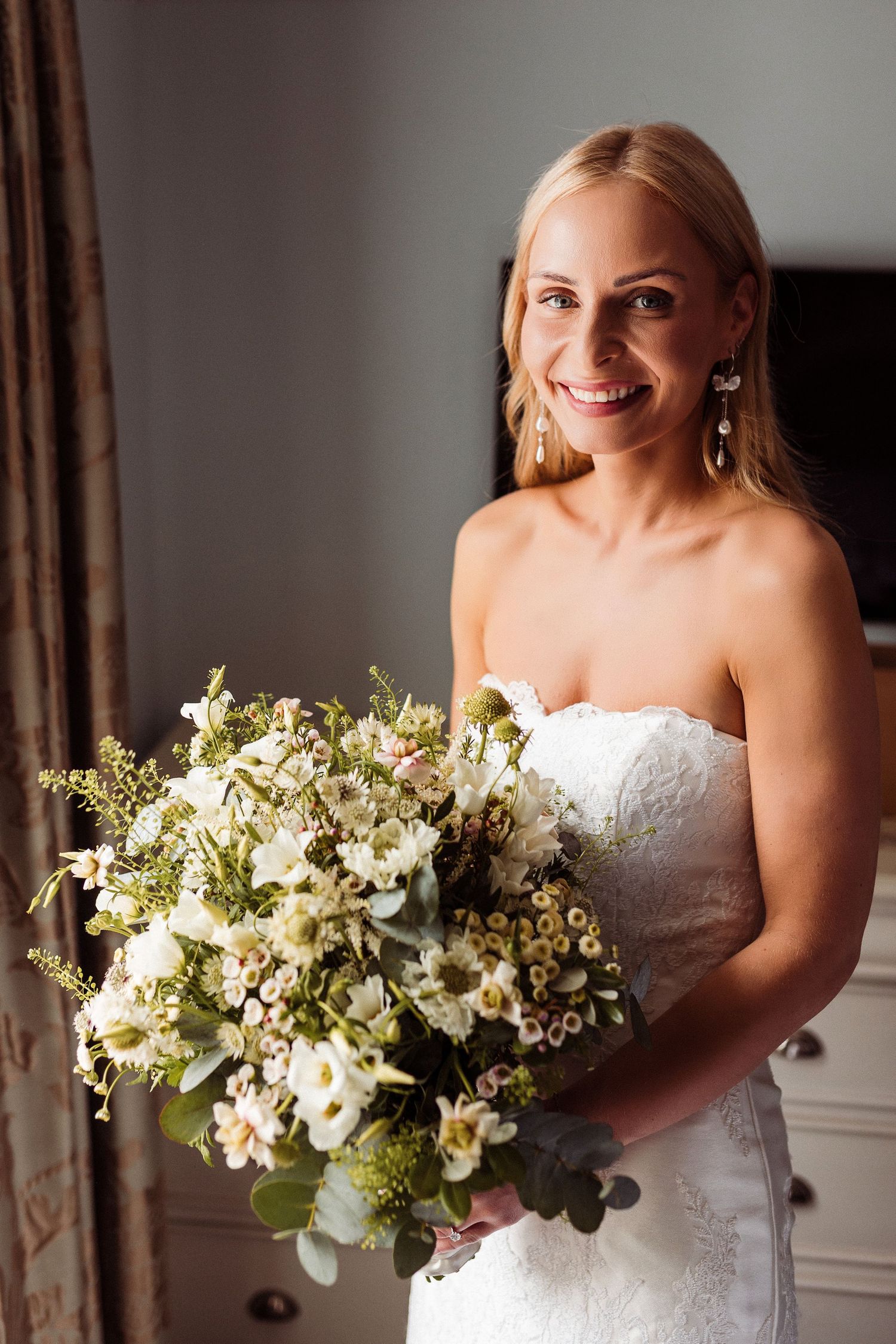 A bride in a strapless white wedding dress holds a lush bouquet of white flowers and greenery against a neutral background.