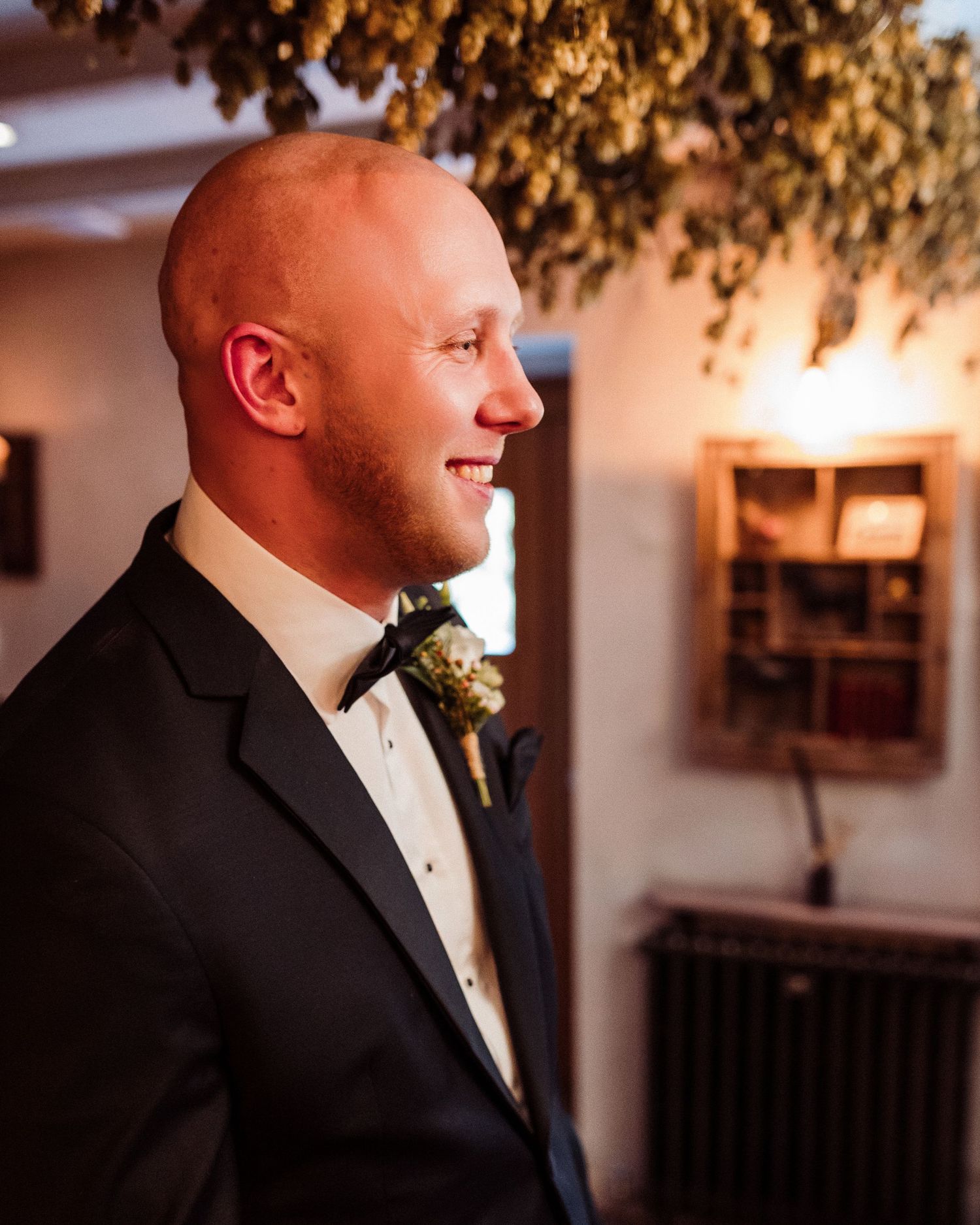 A groom in a black tuxedo and bowtie smiles during a wedding ceremony with warm lighting and decor in the background.