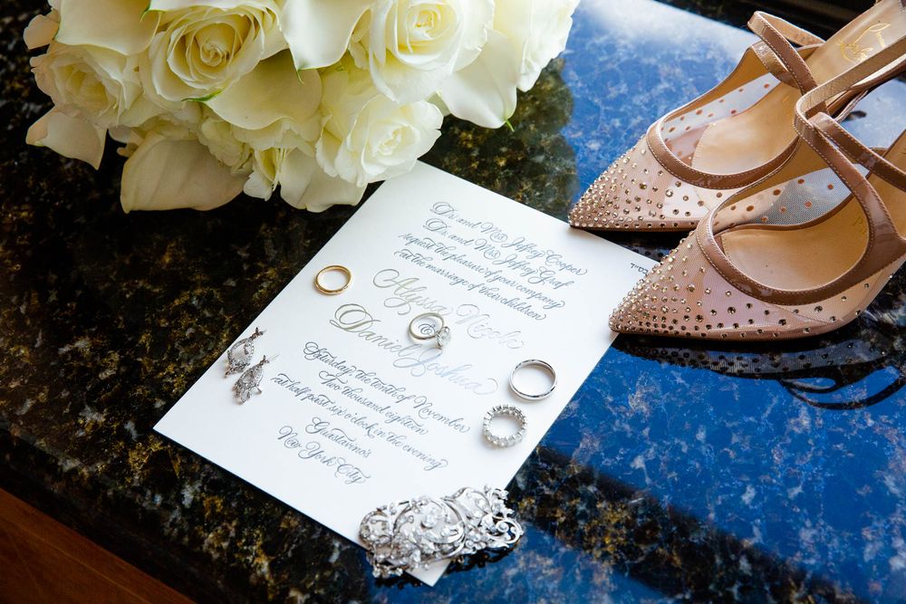 Wedding invitation with rings and bridal shoes arranged on blue surface with white roses.