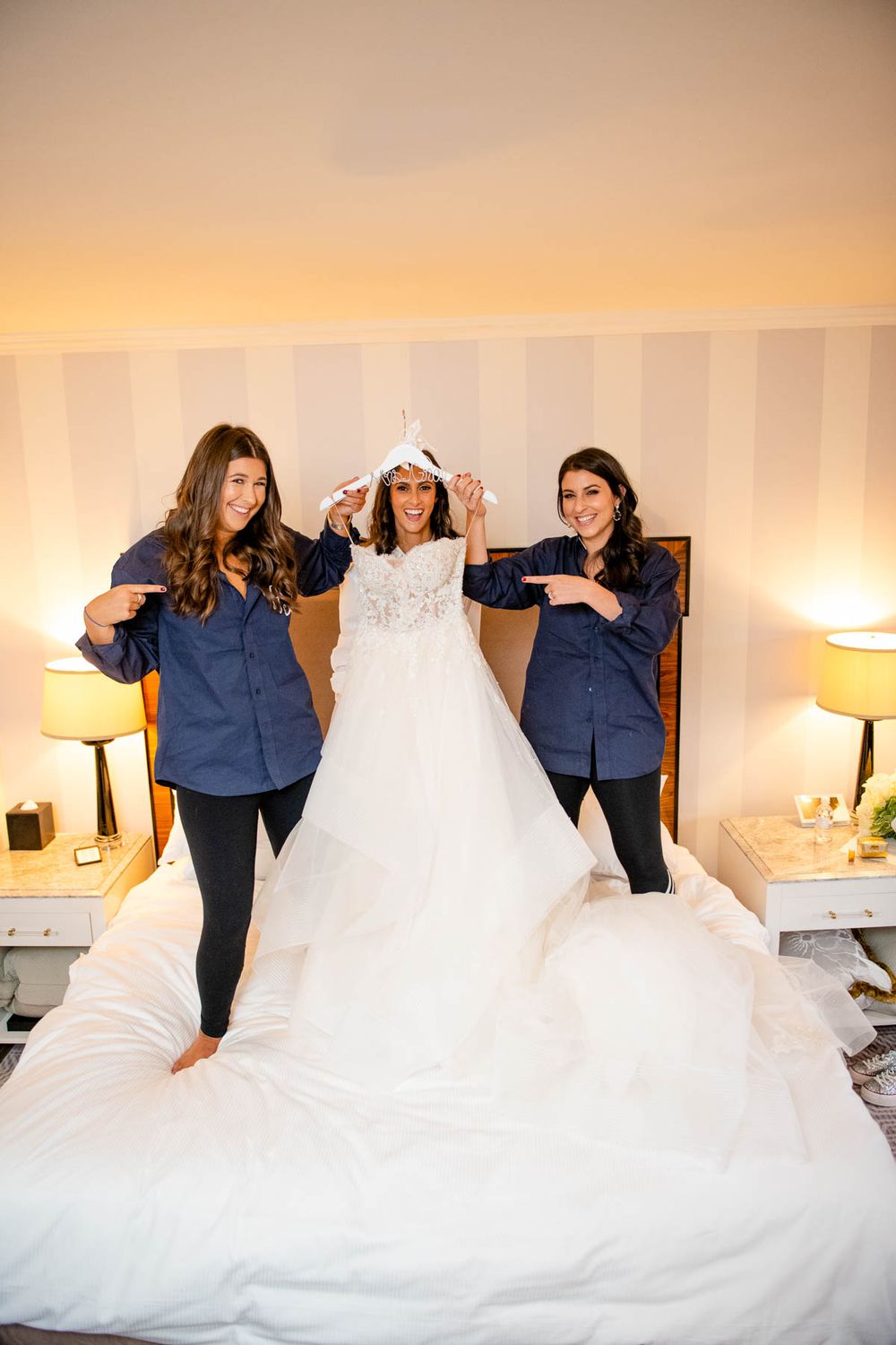 Bridesmaids in navy helping adjust a flowing white wedding gown on bed.