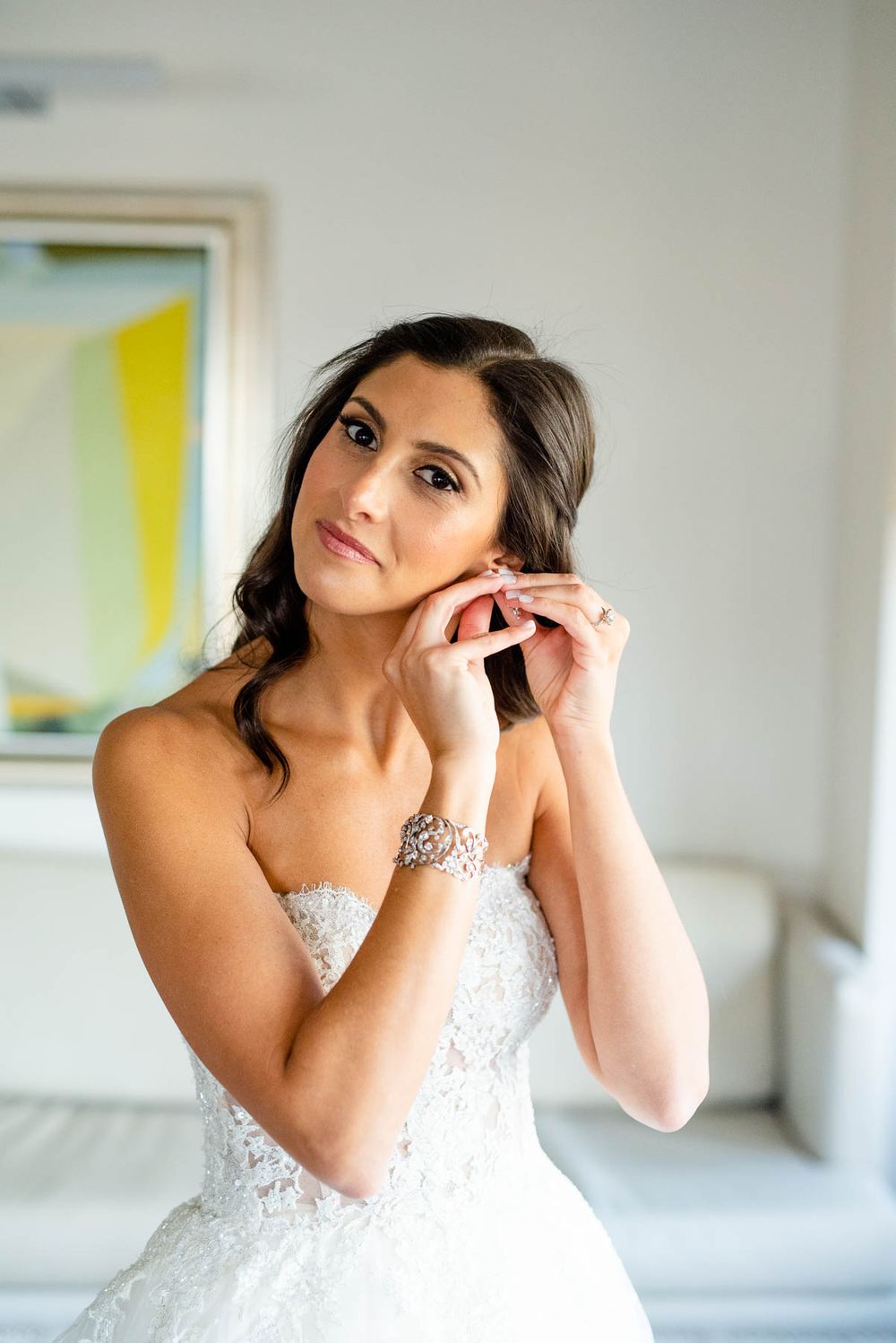 Bride putting on jewelry while wearing strapless lace wedding gown.