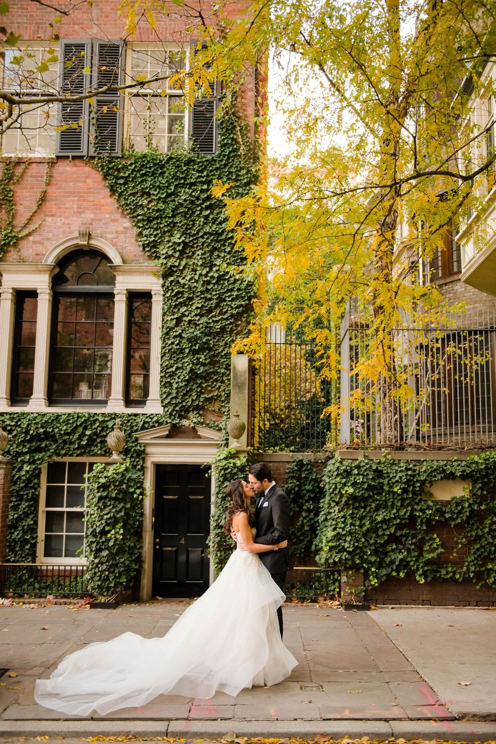 Wedding couple embracing in front of ivy-covered brownstone building during fall season.