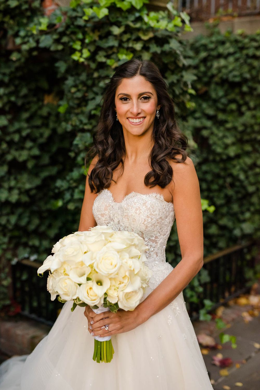 Beautiful bride in strapless lace gown holding white rose bouquet against ivy backdrop.