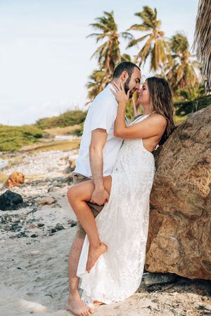 Romantic couple embraces on a tropical beach with palm trees and rocks at sunset.
