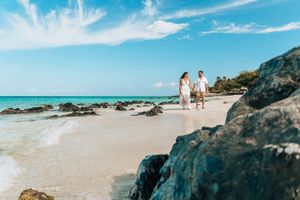 A couple walks along a pristine white sand beach with turquoise waters and rocky cliffs under a bright blue sky.