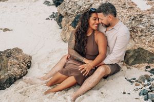 Romantic couple embraces on sandy beach rocks during sunset photoshoot session.
