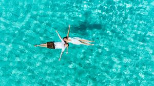 A person floats peacefully in crystal clear turquoise waters while wearing a white swimsuit and hat.