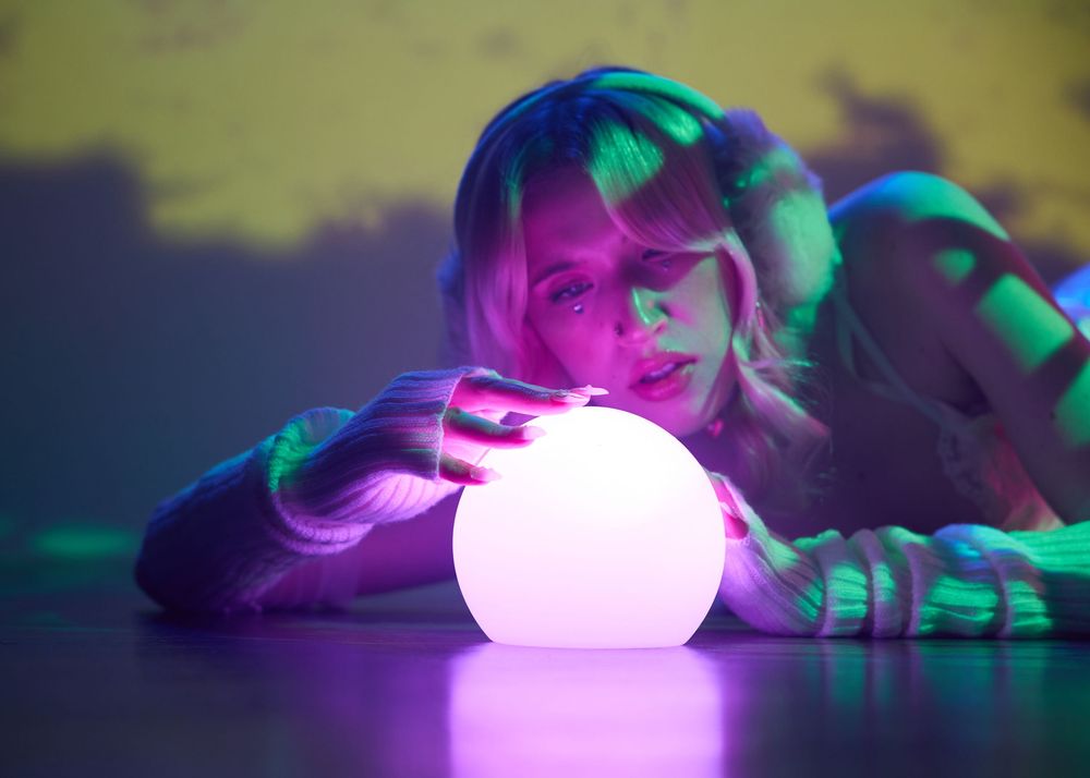 Blonde woman gazing at glowing white orb on reflective surface in neon light.