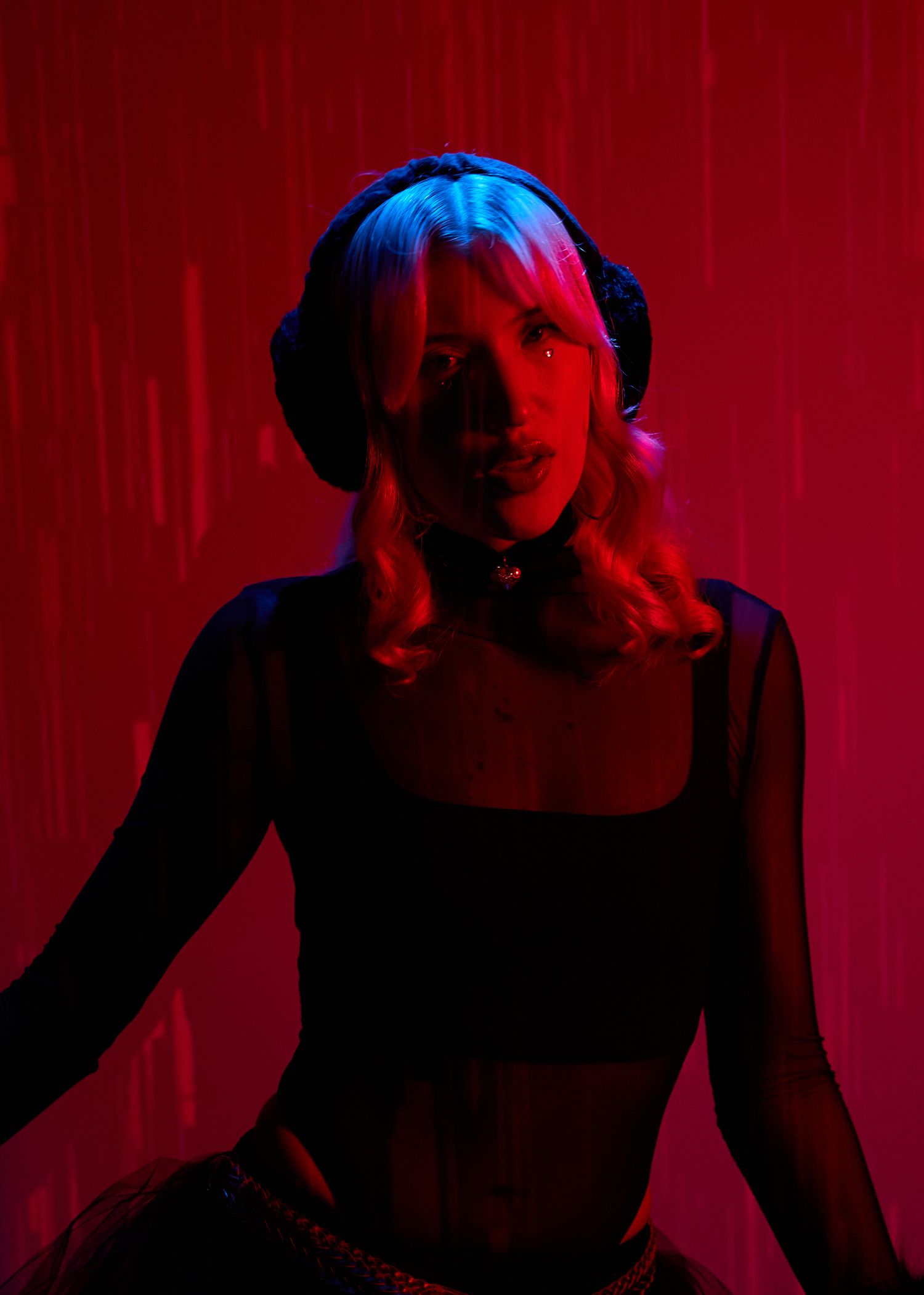 Woman in black crop top and headphones poses under dramatic red and blue neon lighting in a dark studio setting.