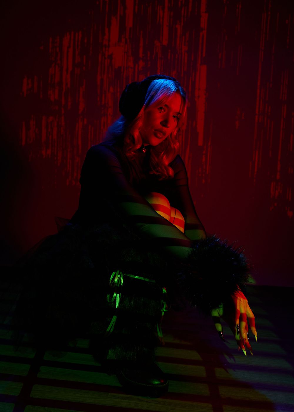 A woman poses dramatically under red and blue neon lights, wearing dark clothing with fur accents and long dark nails.