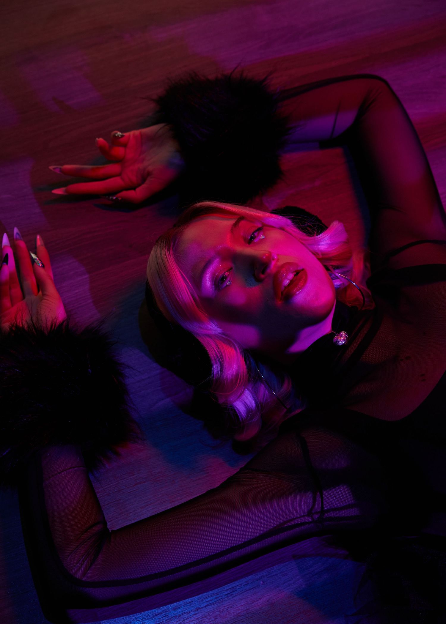 Woman with blonde hair and red nails lying on floor under dramatic pink and purple neon lighting.