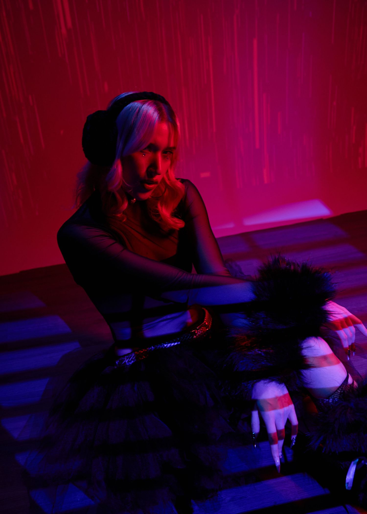Blonde woman with headphones sits in red neon-lit room with checkered floor and stuffed toys nearby.