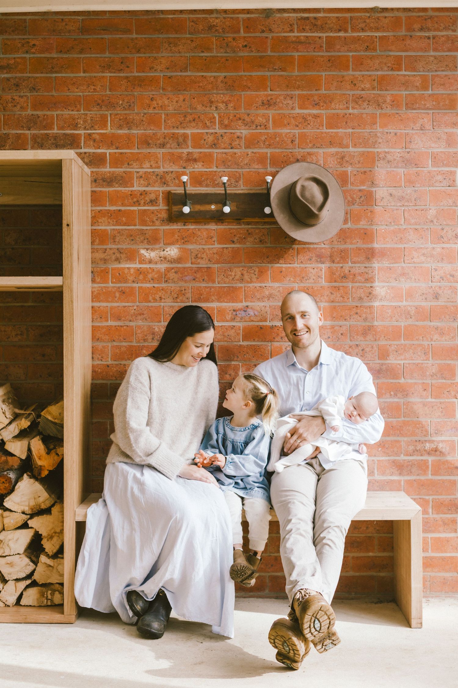 Family of four sitting on a wooden bench against a red brick wall, with stacked firewood and a hat on hooks nearby.