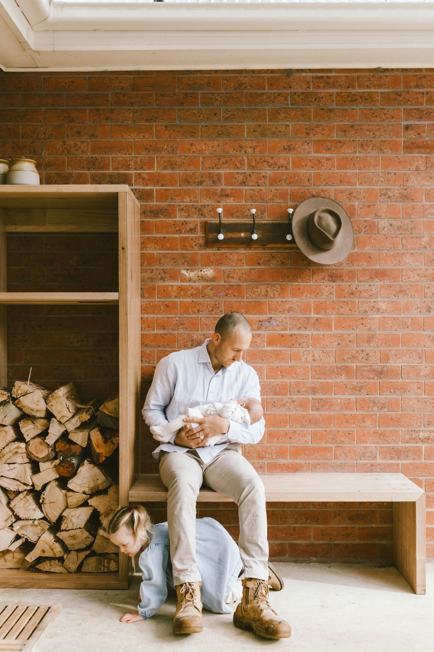 Man sitting on a wooden bench holding a newborn baby against a rustic brick wall with firewood stacked nearby.