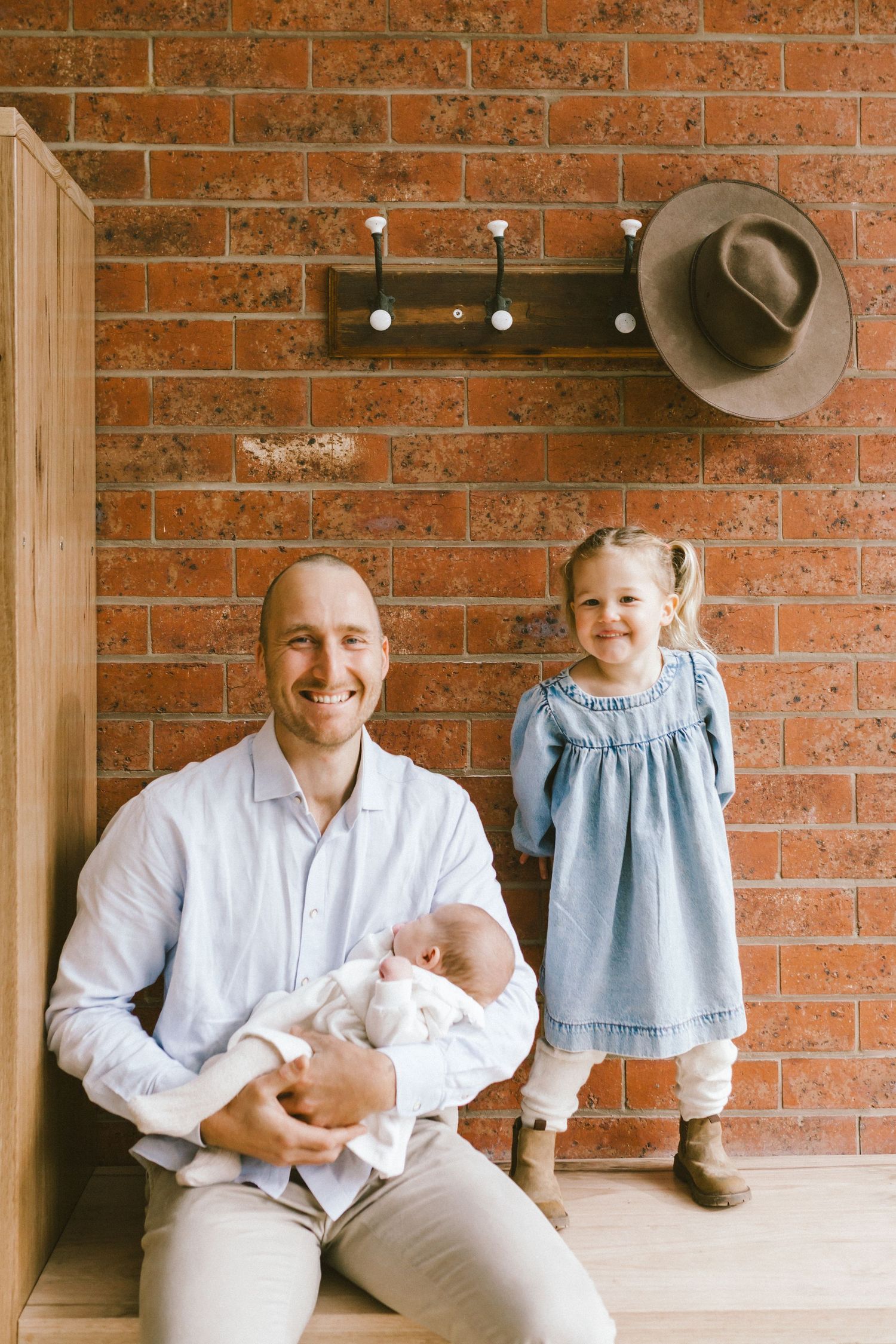 A smiling father holds a newborn while his young daughter in a blue dress stands beside him against a brick wall.