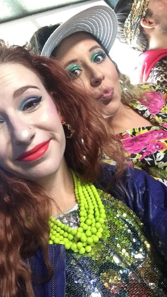 Two women with bold colorful makeup and neon accessories pose for a fun selfie at a festive event.