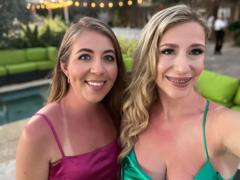 Two smiling women in elegant satin gowns posing together at an outdoor evening event near a pool.