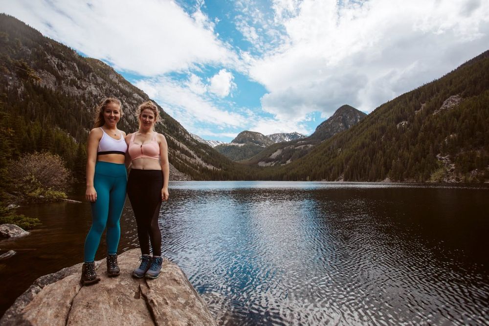 Two women in athletic wear pose on a rock by a serene mountain lake surrounded by forested peaks.