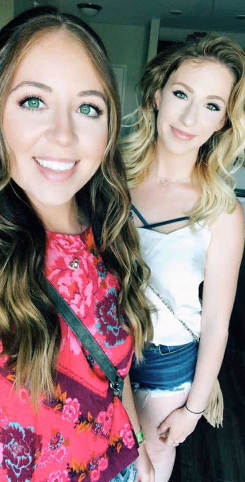 Two smiling young women pose for a selfie, one in a floral top and the other in a white cami with denim shorts.